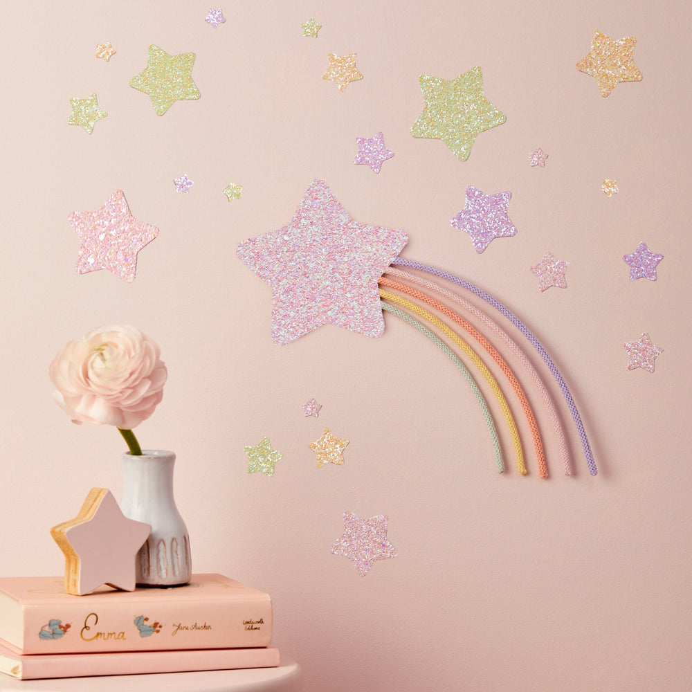 a pink wall with a rainbow-colored star decoration and scattered glitter stars. On a small table, there is a vase with a pink flower and a book titled "Emma".