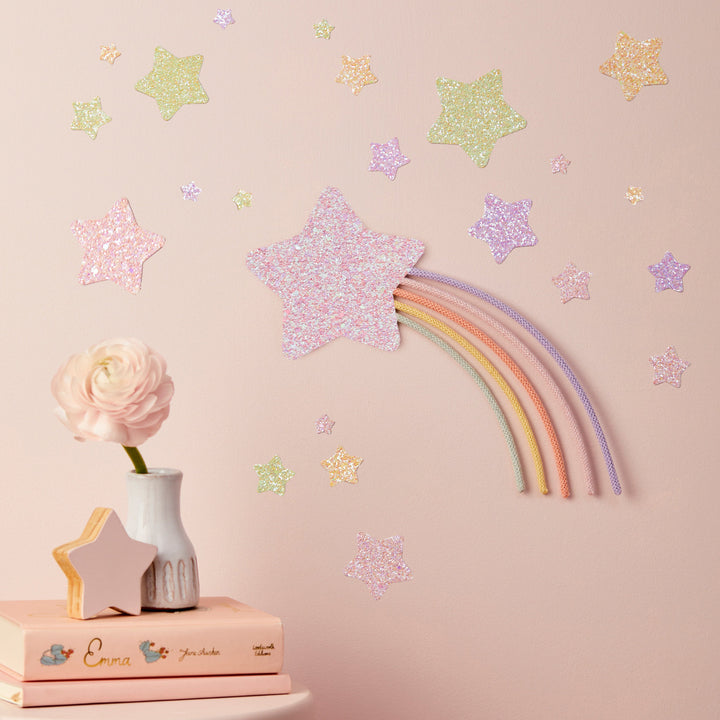 a pink wall with a rainbow-colored star decoration and scattered glitter stars. On a small table, there is a vase with a pink flower and a book titled "Emma".