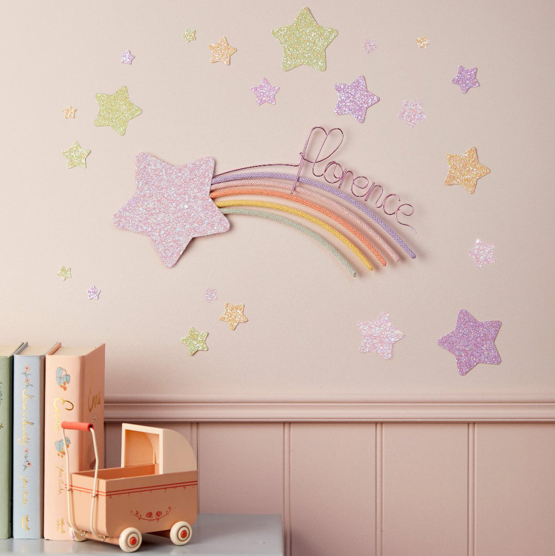 a pink wall decorated with a glittery star and a rainbow-colored wire decoration, along with scattered small star-shaped decorations. There is a pink toy car and a stack of books on a shelf below the wall.