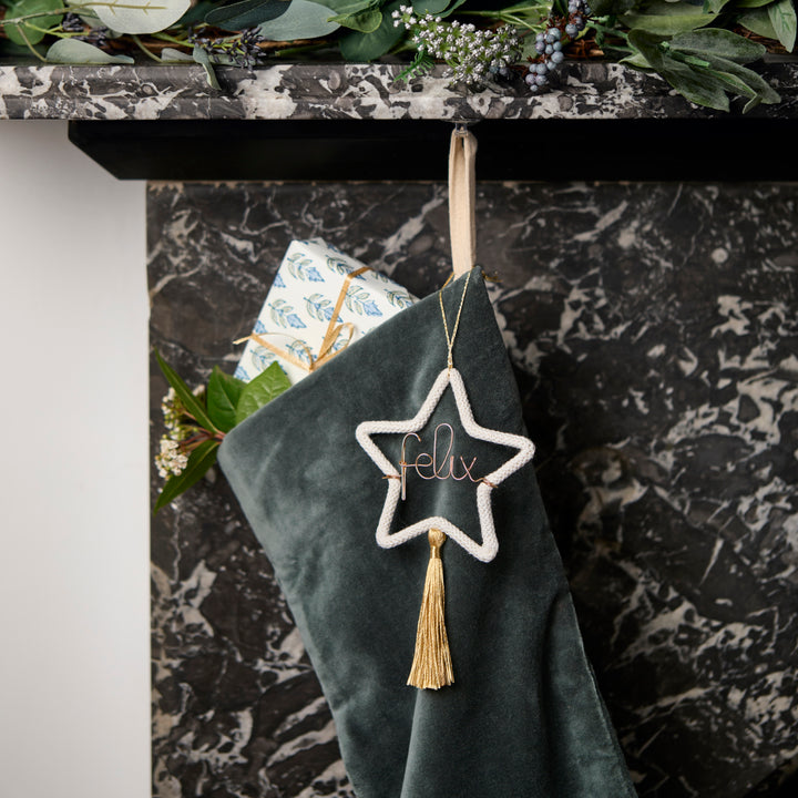Green velvet stocking with personalised star-shaped ornament and tassel hanging on a marble fireplace.