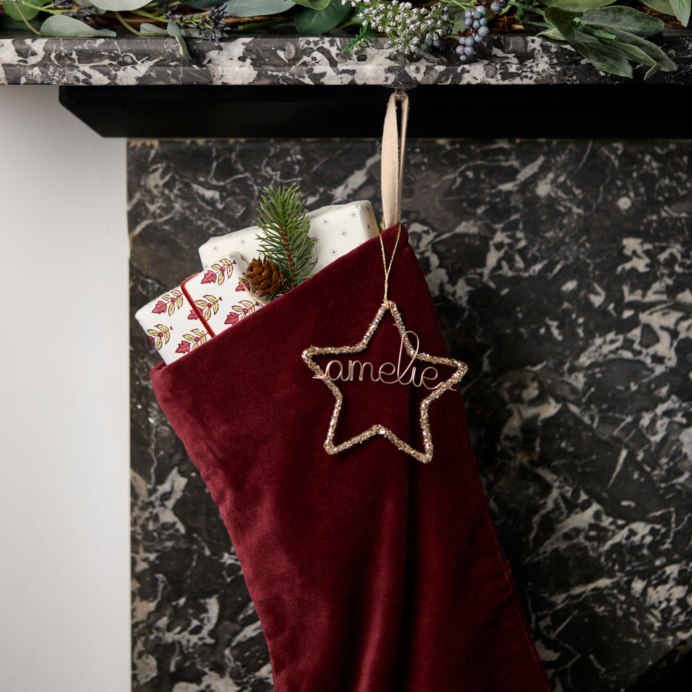 Red Christmas stocking with a gold glitter star personalised ornament hanging on a marble fireplace.