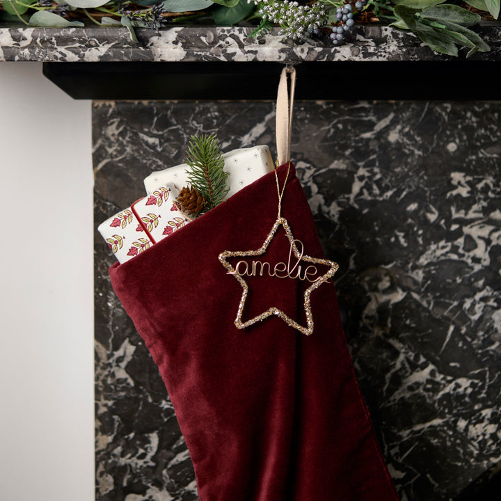 Red Christmas stocking with a gold glitter star personalised ornament hanging on a marble fireplace.