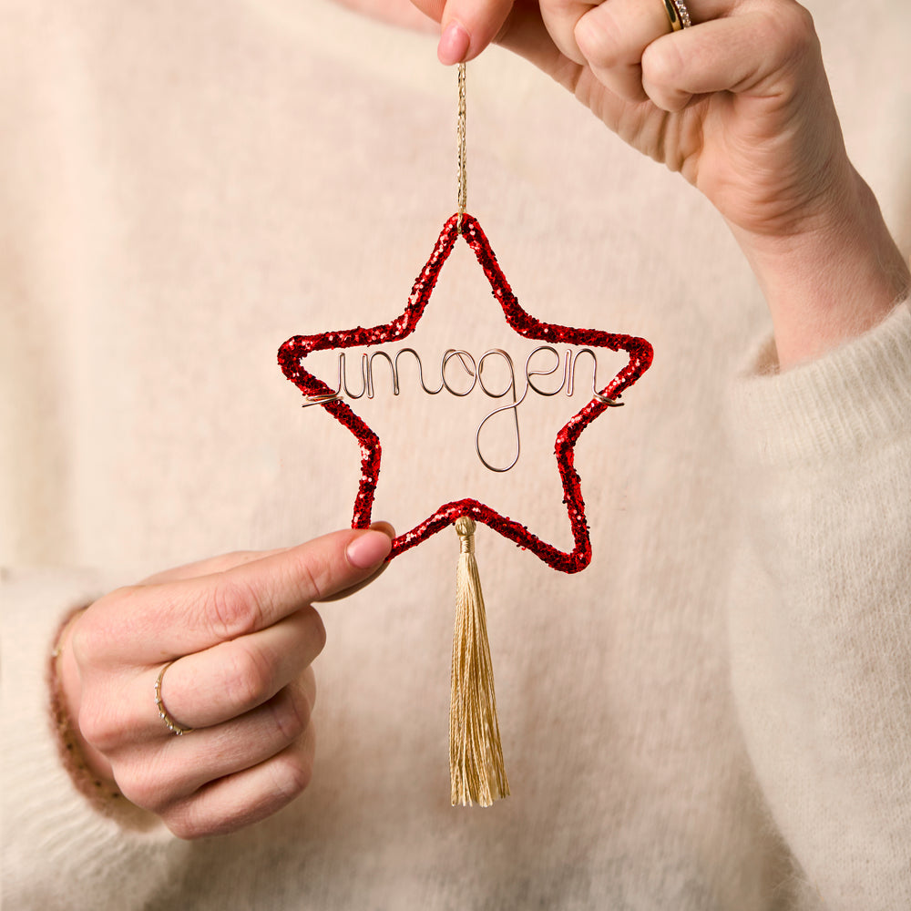 Star-shaped red glitter ornament personalised with 'imogen' name and gold tassel held by a person wearing a beige sweater.