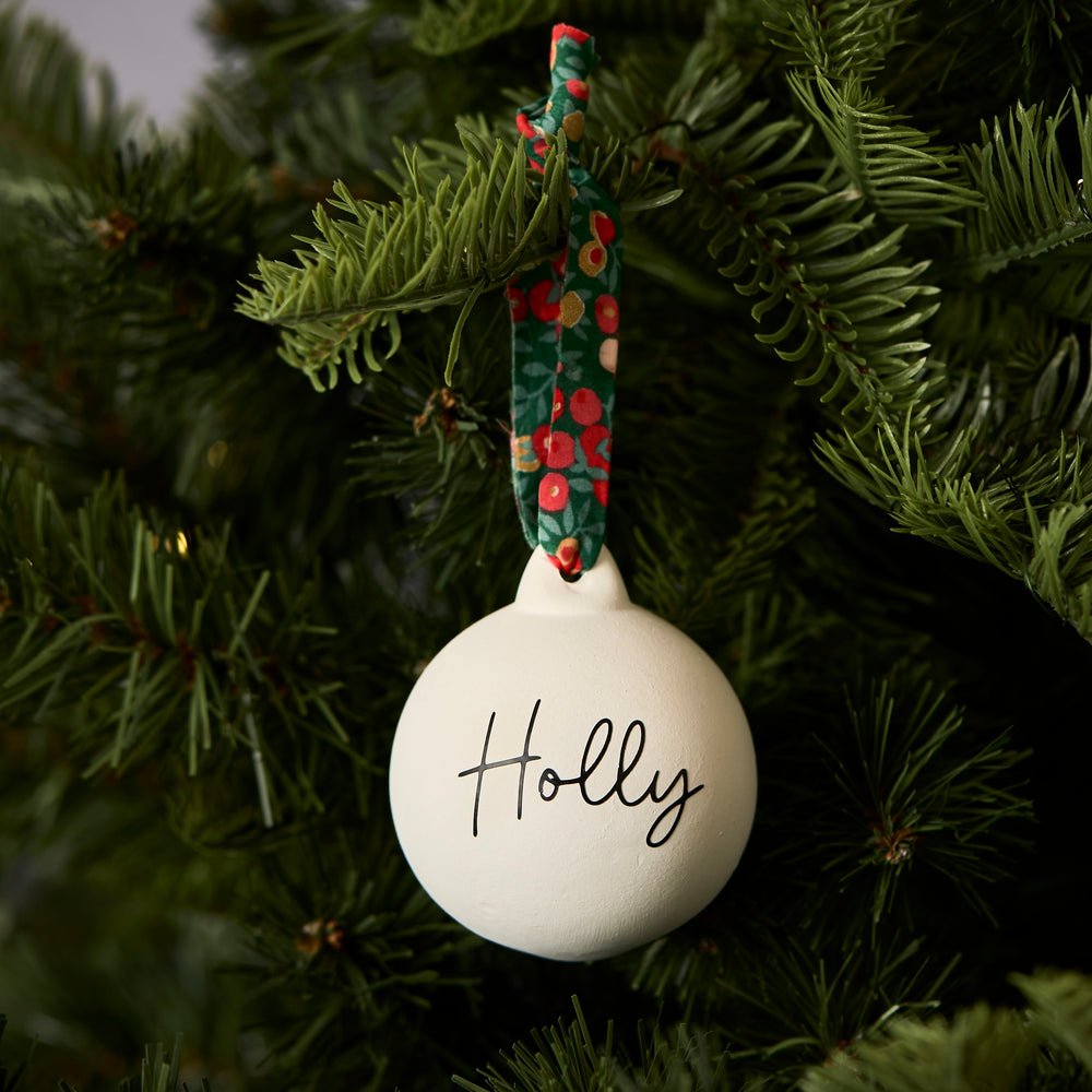 White ceramic bauble personalised with 'Holly' text and tied with floral Liberty London ribbon, hanging on a Christmas tree branch.