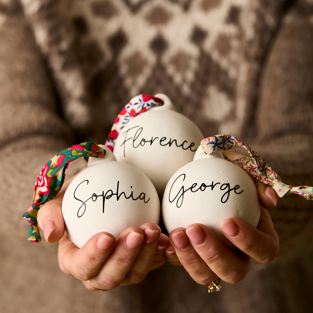 Three ceramic baubles with Liberty London ribbon and personalised  with names 'Sophia', 'Florence', and 'George' held by a person.