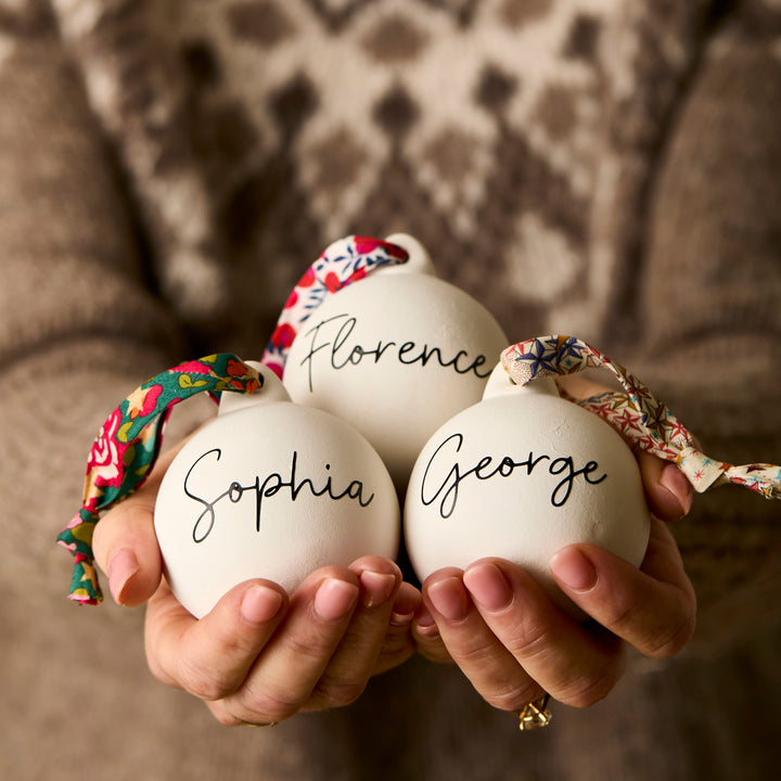 Three ceramic baubles with Liberty London ribbon and personalised  with names 'Sophia', 'Florence', and 'George' held by a person.