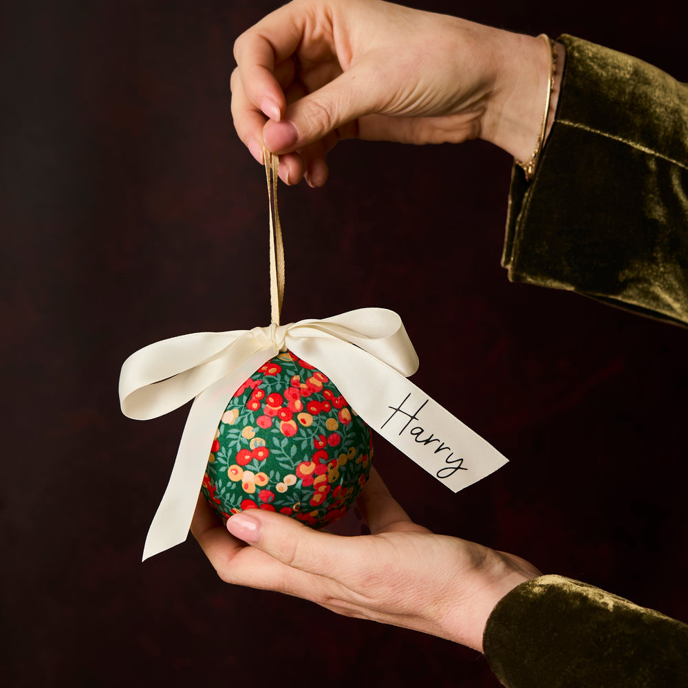 Hand holding a Wiltshire green and gold Liberty London  ornament with a white personalised ribbon, against a dark background.