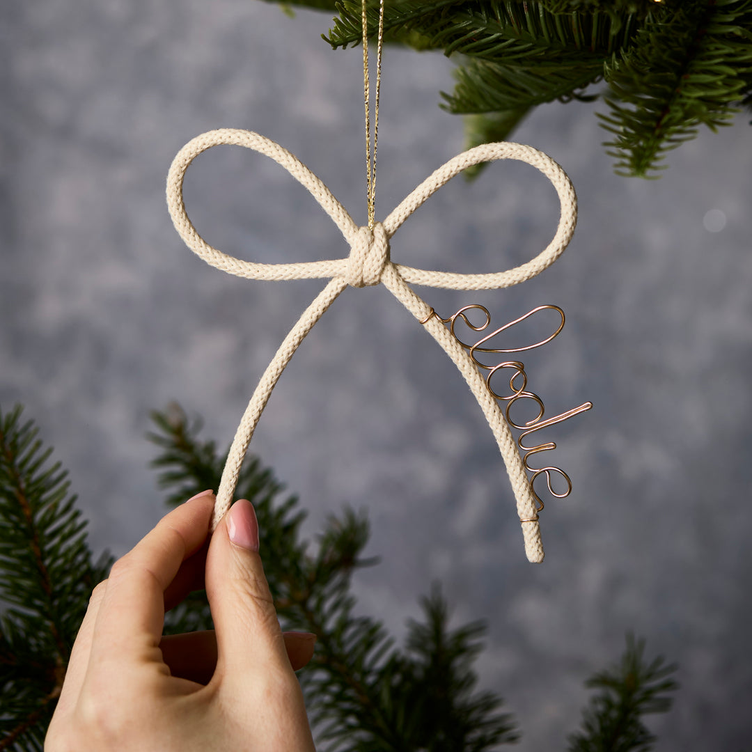 Hand holding a Christmas white knit bow ornament with personalised with name  'Elodie' on a blurred natural background