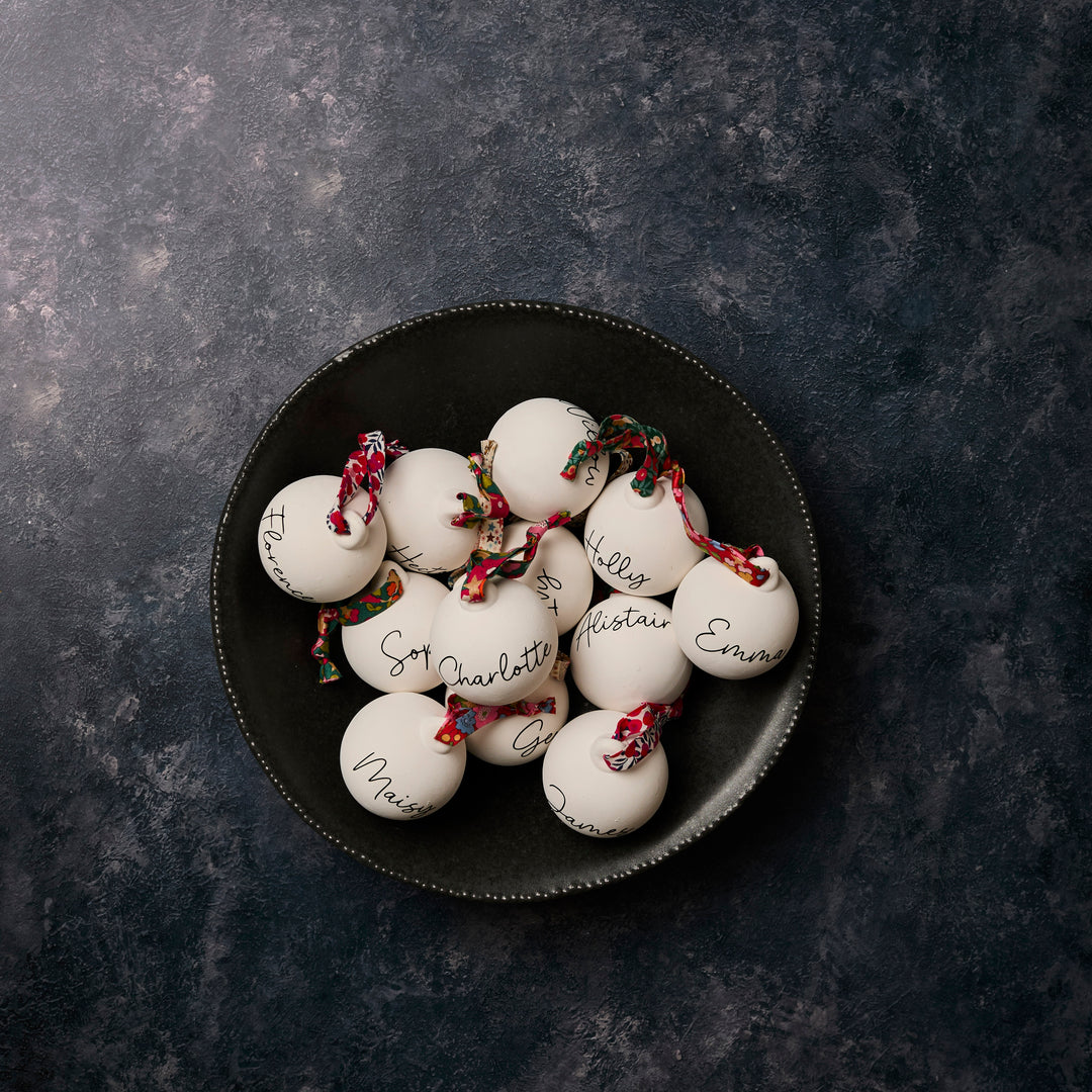 White ceramic ornaments with names written on them, tied with floral Liberty London ribbon, on a dark surface.