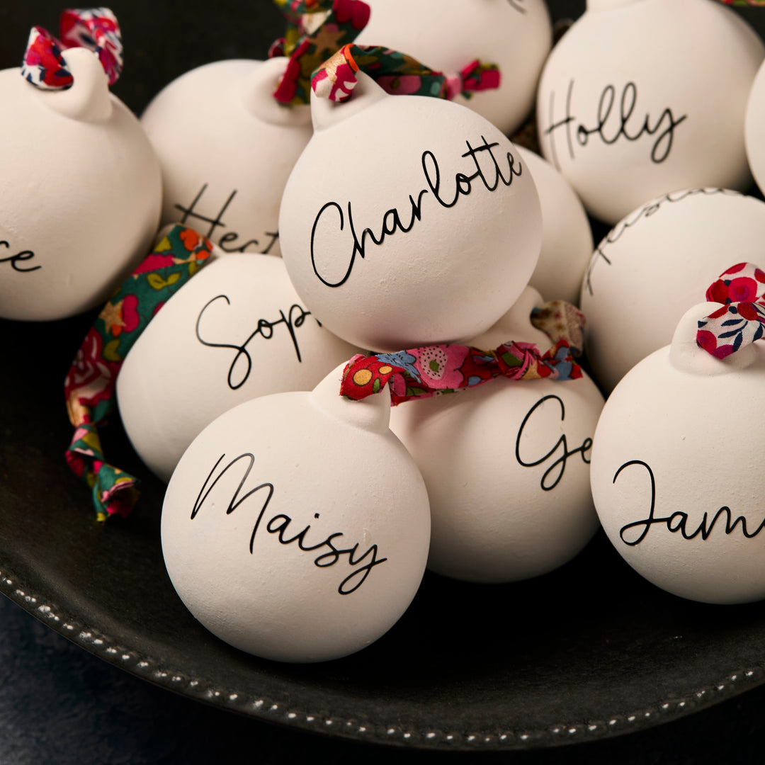 White ceramic ornaments with names written on them, tied with floral Liberty London ribbon, on a dark surface.