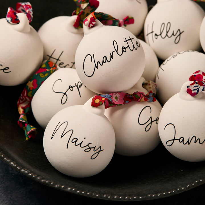 White ceramic ornaments with names written on them, tied with floral Liberty London ribbon, on a dark surface.