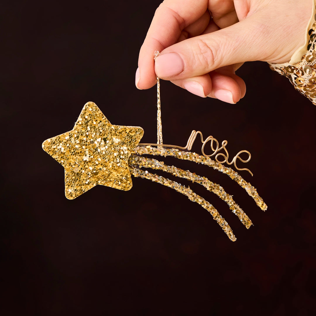 A hand wearing a gold lace sleeve holds a gold glittery shooting star ornament with the word 'rose' in cursive script against a dark brown background.